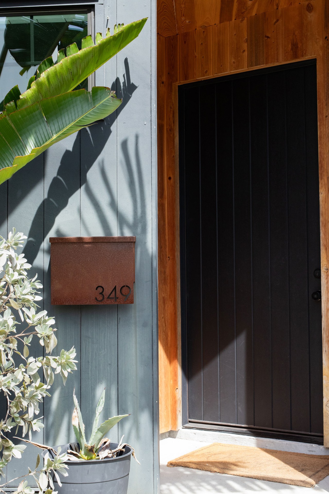 Pioneer Built entryway — black vertical-plank front door with cedar surround, a rust mailbox marked 349, and tropical plantings